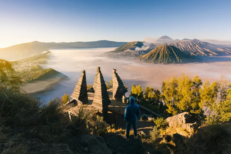 Seseorang berdiri di teras pandang Penanjakan menghadap gapura kayu, dengan Bromo, Batok, dan Semeru di seberang kaldera yang tertutup awan.